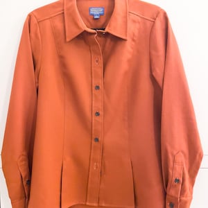 以下が含まれることがあります： オレンジ色のボタンダウンシャツ。襟と長袖が付いています。ウエストにはプリーツのデザインがあり、襟には小さなブランドラベルが付いています。ボタンは濃い茶色です。シャツは黒いハンガーにかかっています。