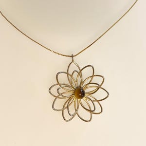 May include: A gold-toned necklace featuring a pendant shaped like a stylized flower with multiple thin, open wire petals radiating from a central, round, brown tiger's eye gemstone. The pendant hangs from a delicate, matching gold-toned chain.