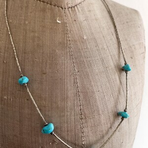 May include: A silver necklace featuring five turquoise-colored stones. The stones are irregularly shaped and spaced along the necklace's silver chain. The necklace is displayed on a mannequin.