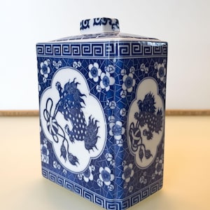 Peut inclure: Un récipient en céramique bleu et blanc de forme carrée avec un couvercle assorti. Le récipient présente un motif floral répété et un motif central d'une créature mythique. Le couvercle a un petit bouton décoratif.