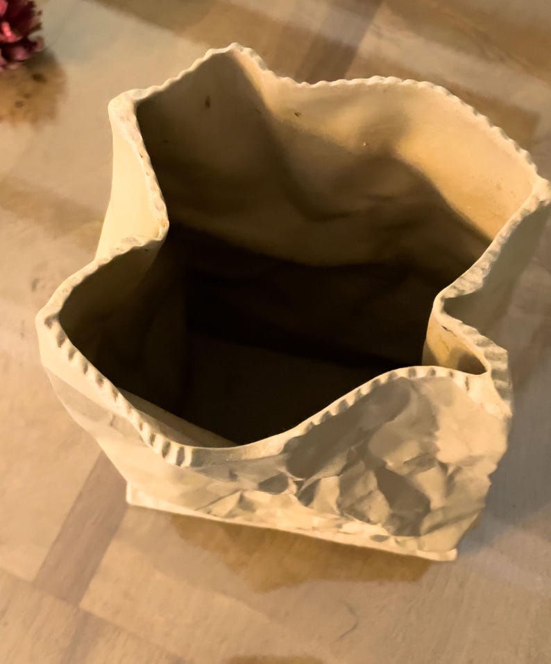 May include: A beige ceramic vase designed to resemble a crumpled paper bag. The vase has a wide, irregular opening and a textured surface, with visible creases and folds. The interior is a darker shade of brown.