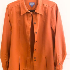 Può includere: Una camicia arancione bruciato con bottoni, maniche lunghe e colletto. La camicia ha una vestibilità su misura con cuciture verticali e una piccola etichetta con la scritta "Pendleton". La camicia è appesa a una gruccia nera.