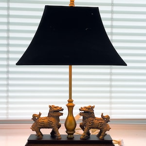 May include: A table lamp featuring a black rectangular fabric shade and a gold-toned metal central stem. The base consists of a dark rectangular wooden platform adorned with two gold-colored mythical lion figures facing each other.