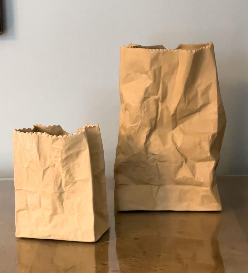 May include: Two brown paper bags of different sizes stand side-by-side. The bags have a crumpled texture and open, jagged tops. The bags are on a reflective surface, and the background is a solid, light blue-gray.