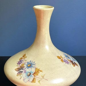 Puede incluir: Un jarrón de cerámica de color crema con acabado moteado, base ancha y redondeada que se estrecha hacia un cuello fino. El cuerpo del jarrón está decorado con dos motivos florales que presentan pétalos azules, morados y hojas marrones.