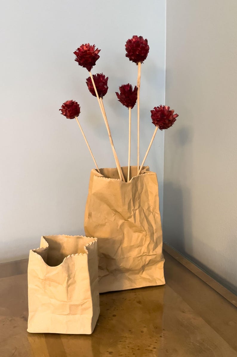 May include: Two brown paper bags, one large and one small, sit on a reflective surface. The larger bag holds several dried, burgundy-colored flower stems. The background is a light blue wall.
