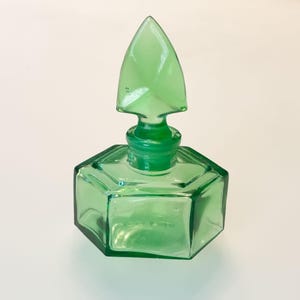 Op de afbeelding: Een doorschijnend smaragdgroen glazen parfumflesje met een zeshoekige basis en een bijpassende puntige, gefacetteerde glazen stop. Het flesje heeft een korte, smalle hals en platte, hoekige zijkanten die een geometrisch silhouet vormen.