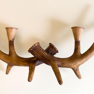 Puede incluir: Un candelabro decorativo con forma de cuernos de ciervo, con dos portavelas. El soporte está hecho de un material marrón claro, con los cuernos cruzándose en el centro. Los portavelas son de un marrón más claro. Un artículo de decoración único.