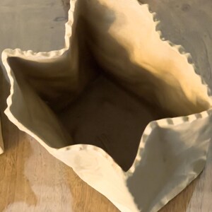 May include: A beige ceramic vase with a unique, folded paper bag design. The vase has a textured surface and a jagged, uneven rim. The interior is a darker shade of beige, creating a contrast with the exterior. The vase sits on a wooden surface.