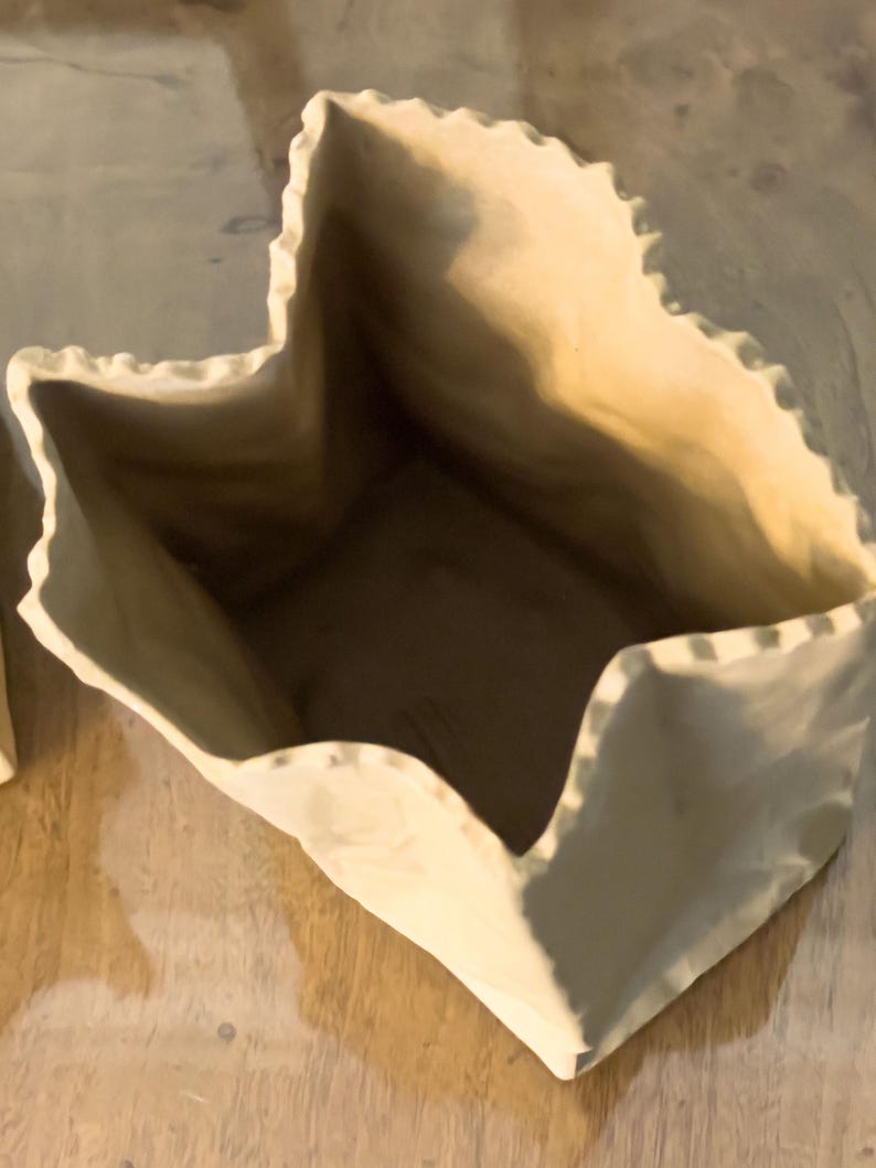 May include: A beige ceramic vase with a unique, folded paper bag design. The vase has a textured surface and a jagged, uneven rim. The interior is a darker shade of beige, creating a contrast with the exterior. The vase sits on a wooden surface.