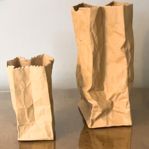 May include: Two brown paper bag sculptures with a crinkled texture. One bag is tall and upright, while the other is shorter and tilted. The edges of the bags have a decorative, jagged cut. The bags are made of a light brown paper.