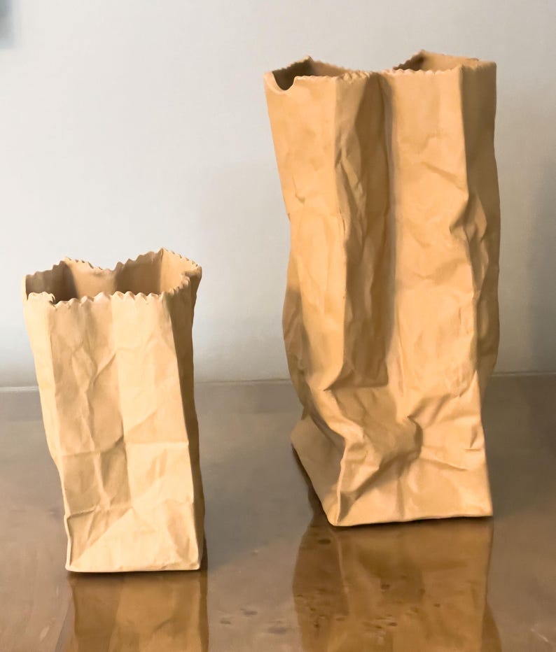 May include: Two brown paper bag sculptures with a crinkled texture. One bag is tall and upright, while the other is shorter and tilted. The edges of the bags have a decorative, jagged cut. The bags are made of a light brown paper.