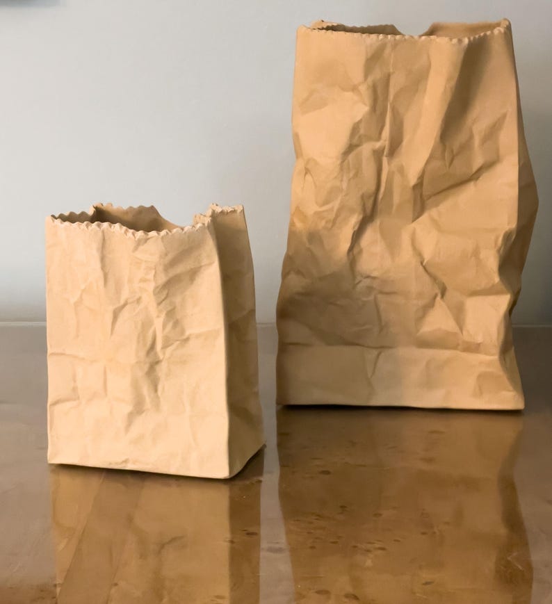 May include: Two brown paper bags of different sizes stand side-by-side. The bags have a crumpled texture and a rectangular base. The edges of the bags are uneven, giving them a handmade appearance. The bags are on a reflective surface.