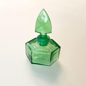 Op de afbeelding: Een doorschijnend smaragdgroen glazen parfumflesje met een zeshoekige basis en een bijpassende puntige, speervormige glazen stop. Het object heeft strakke, geometrische lijnen en een glad, gepolijst oppervlak dat licht door de groene tint weerkaatst.