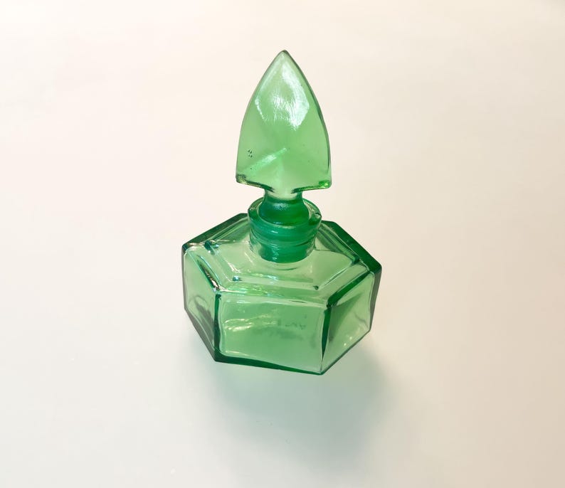 Op de afbeelding: Een doorschijnend smaragdgroen glazen parfumflesje met een zeshoekige basis en een bijpassende puntige, speervormige glazen stop. Het object heeft strakke, geometrische lijnen en een glad, gepolijst oppervlak dat licht door de groene tint weerkaatst.