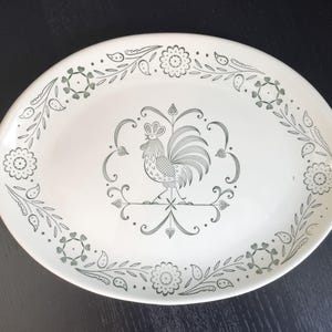 May include: An oval-shaped white ceramic serving platter featuring a dark green line-art illustration of a rooster centered within a decorative frame. The rim is adorned with a repeating pattern of stylized flowers, leaves, and paisley shapes.