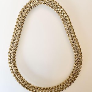 May include: A gold-toned chain necklace with a double-strand design. The necklace features a series of interlocking links, creating a textured and bold appearance. The clasp is visible at the top, securing the necklace.