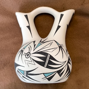 May include: White ceramic vase with a handle and two spouts. The vase features a black and turquoise geometric design. The design includes lightning bolt shapes and stylized floral elements. The vase is likely handmade.