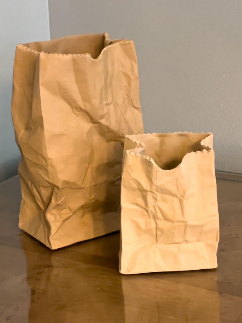 May include: Two brown paper bags of different sizes, with crumpled textures and open tops. The bags are standing on a reflective surface, with a neutral-colored wall in the background. The bags have a classic, simple design.