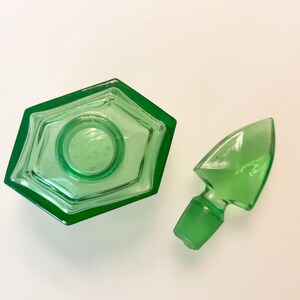 Op de afbeelding: Een doorschijnend smaragdgroen glazen parfumflesje met een zeshoekige basis en een bijpassende puntige glazen stop. Het flesje heeft een centrale ronde opening en gefacetteerde randen, met de stop los naast het flesje geplaatst.