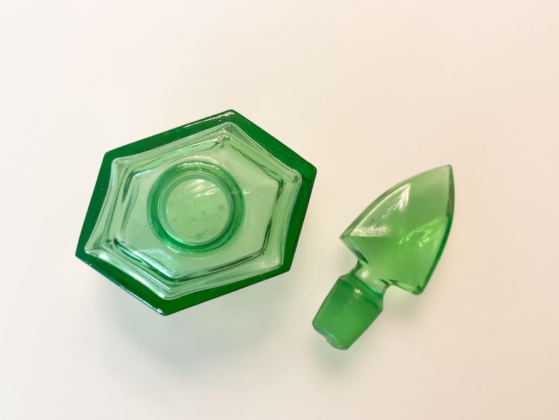 Op de afbeelding: Een doorschijnend smaragdgroen glazen parfumflesje met een zeshoekige basis en een bijpassende puntige glazen stop. Het flesje heeft een centrale ronde opening en gefacetteerde randen, met de stop los naast het flesje geplaatst.