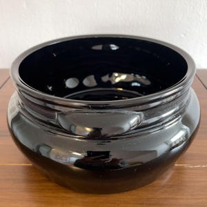 Può includere: Una ciotola in ceramica nera lucida, di forma ampia e arrotondata. Presenta una fascia strutturata al centro e piccole maniglie. L'interno è liscio e riflettente. Questa ciotola decorativa è adatta a vari usi.