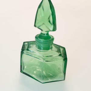 Op de afbeelding: Een doorschijnend lichtgroen glazen parfumflesje met een zeshoekige basis en een bijpassende puntige, gefacetteerde glazen stop. Het flesje heeft strakke, geometrische lijnen en een glad, gepolijst oppervlak dat de levendige groene kleur toont.