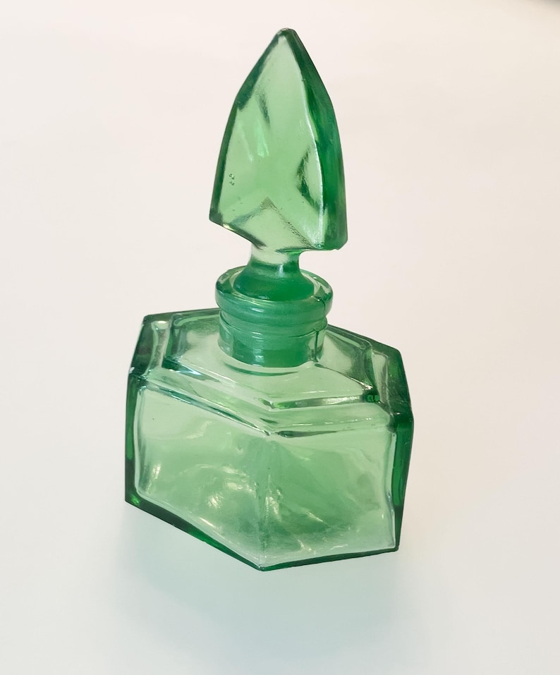 Op de afbeelding: Een doorschijnend lichtgroen glazen parfumflesje met een zeshoekige basis en een bijpassende puntige, gefacetteerde glazen stop. Het flesje heeft strakke, geometrische lijnen en een glad, gepolijst oppervlak dat de levendige groene kleur toont.