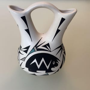 May include: White ceramic vase with a handle and two spouts. The vase is decorated with black geometric shapes and a stylized black and white turtle design. Accents of teal are also present.