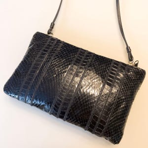 May include: A black rectangular crossbody bag with a textured, reptile-like pattern. The bag has a long, thin strap and a zipper closure. The bag is made of a dark, shiny material.