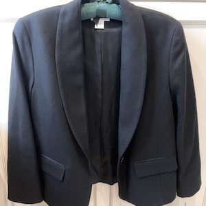 Può includere: Un blazer nero con ampi revers e due tasche frontali è appeso a un'appendiabiti. Il blazer è realizzato in un tessuto scuro e tinta unita e ha una chiusura con un solo bottone. La fodera interna è nera.