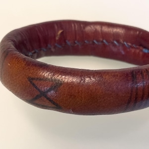 May include: A brown leather bracelet with a stitched seam and a black cross design. The bracelet has a worn appearance and a slightly curved shape. The leather has a reddish-brown hue.