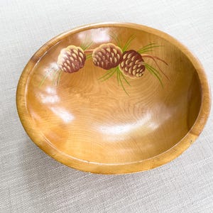 May include: A round, wooden bowl with a glossy finish. The bowl features a hand-painted design of three brown pine cones with green needles. The bowl is a light brown color and sits on a neutral-colored surface.