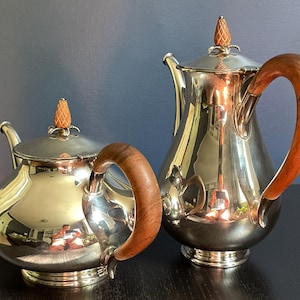 May include: A silver-plated teapot and a taller silver-plated coffee pot, both featuring polished metallic bodies, curved wooden handles, and lids topped with small, textured, light-colored finials shaped like pinecones or decorative buds.