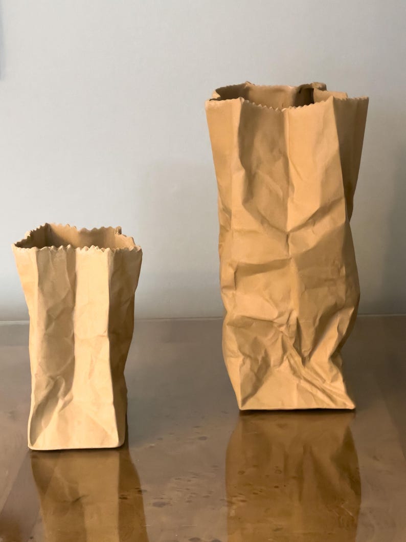 May include: Two brown paper bags of different sizes, standing upright. The bags have a crinkled texture and a jagged top edge. The bags are on a reflective surface, with a light gray background.