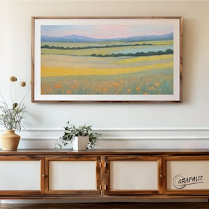 May include: A framed landscape painting in a wooden frame. The artwork shows rolling hills in shades of yellow, green, and blue, with a pink and blue sky. The painting is mounted above a wooden cabinet.