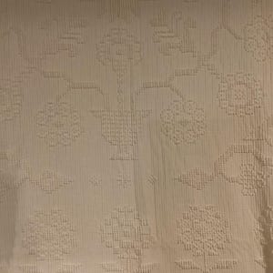 May include: Cream-colored textile with an embossed floral pattern. The design features stylized flowers, leaves, and geometric shapes, creating a textured surface. The fabric appears to be a soft, woven material, suitable for bedding or upholstery.