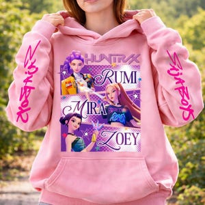May include: Pink hooded sweatshirt with a graphic featuring four animated female characters named Rumi, Mira, and Zoey. The sleeves have the text "Slay Boys" in pink. The sweatshirt has a front pocket.