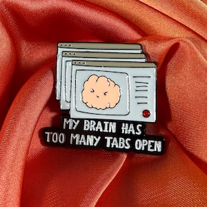 Può includere: Spilla smaltata raffigurante una pila di finestre di computer con un cervello a cartoni animati. La spilla riporta la scritta "MY BRAIN HAS TOO MANY TABS OPEN" in bianco su sfondo nero. La spilla è azzurra, bianca, nera e color pesca.