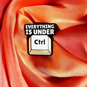 May include: A pin featuring a computer key design with the text "EVERYTHING IS UNDER Ctrl" in black and white. The pin is set against a vibrant orange fabric background.