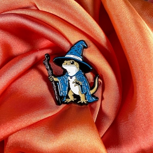 Może przedstawiać: Kapryśna emaliowana przypinka przedstawiająca jaszczurkę ubraną jako czarodziej. Jaszczurka ma na sobie niebieski kapelusz i szatę czarodzieja, trzymając laskę. Przypinka ma czarne kontury i jest umieszczona na żywym pomarańczowym tle.