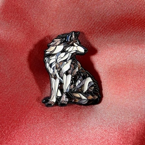 May include: An enamel pin depicting a sitting wolf. The pin features a mosaic design in black, white, and brown. The wolf is set against a red background.
