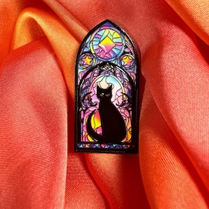 Puede incluir: Pin con silueta de gato negro sobre un diseño de vidriera. El pin presenta un gato negro con cola curvada, en una vidriera colorida con un arcoíris de colores. El pin está sobre una tela rojo-naranja.