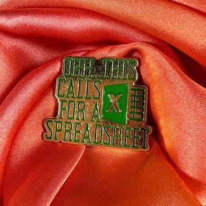 May include: A gold-colored enamel pin with the text "OHH THIS CALLS FOR A SPREADSHEET" in green lettering. The pin features a green Excel logo. The pin is set against a backdrop of orange fabric.