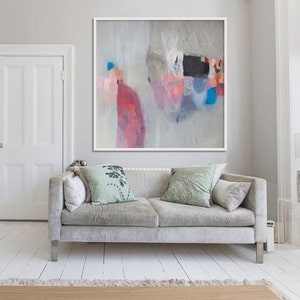 Può includere: Dipinto astratto in cornice bianca. L'opera presenta una miscela di colori, tra cui rosa, rosso, blu e nero, su uno sfondo grigio chiaro. Sotto l'opera c'è un divano grigio con cuscini.
