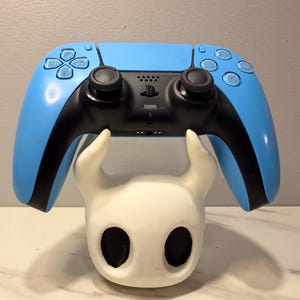 Op de afbeelding: Een lichtblauwe en zwarte PlayStation 5-controller die rust op een witte, gestileerde karakterkop. De controller heeft zwarte joysticks en knoppen. De karakterkop heeft grote, zwarte ovale ogen.