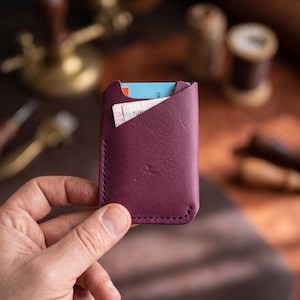 Könnte beinhalten: Ein tiefvioletter Kartenhalter aus Leder mit diagonalem Taschen-Design und sichtbaren dunklen Nähten an den Kanten. Der Kartenhalter enthält eine blaue Karte sowie einen kleinen Stapel Papiergeld im Hauptfach.