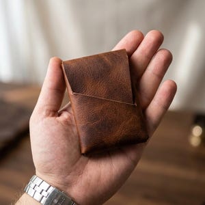 Puede incluir: Un tarjetero de cuero marrón con un acabado texturizado. El tarjetero se sostiene en una mano, mostrando su diseño compacto. El reloj en la muñeca tiene una correa plateada.