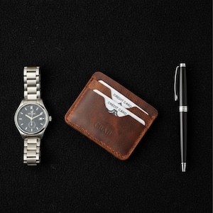 May include: A silver wristwatch with a dark gray face, a brown leather wallet with two credit cards, and a black pen with silver accents are arranged on a black surface. The wallet is embossed with the word "CRAB".