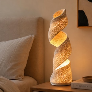 May include: A spiral-shaped table lamp featuring a textured, off-white woven shade that emits a warm, soft glow. The lamp has a continuous, twisting design that rises from a circular base, creating an open, sculptural form that illuminates.